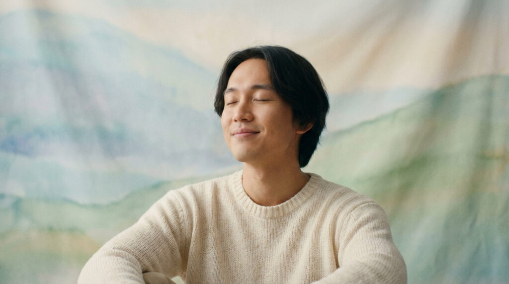 Homme asiatique aux yeux fermés, souriant, en pull beige, méditant pour la gestion de la douleur sur fond abstrait.